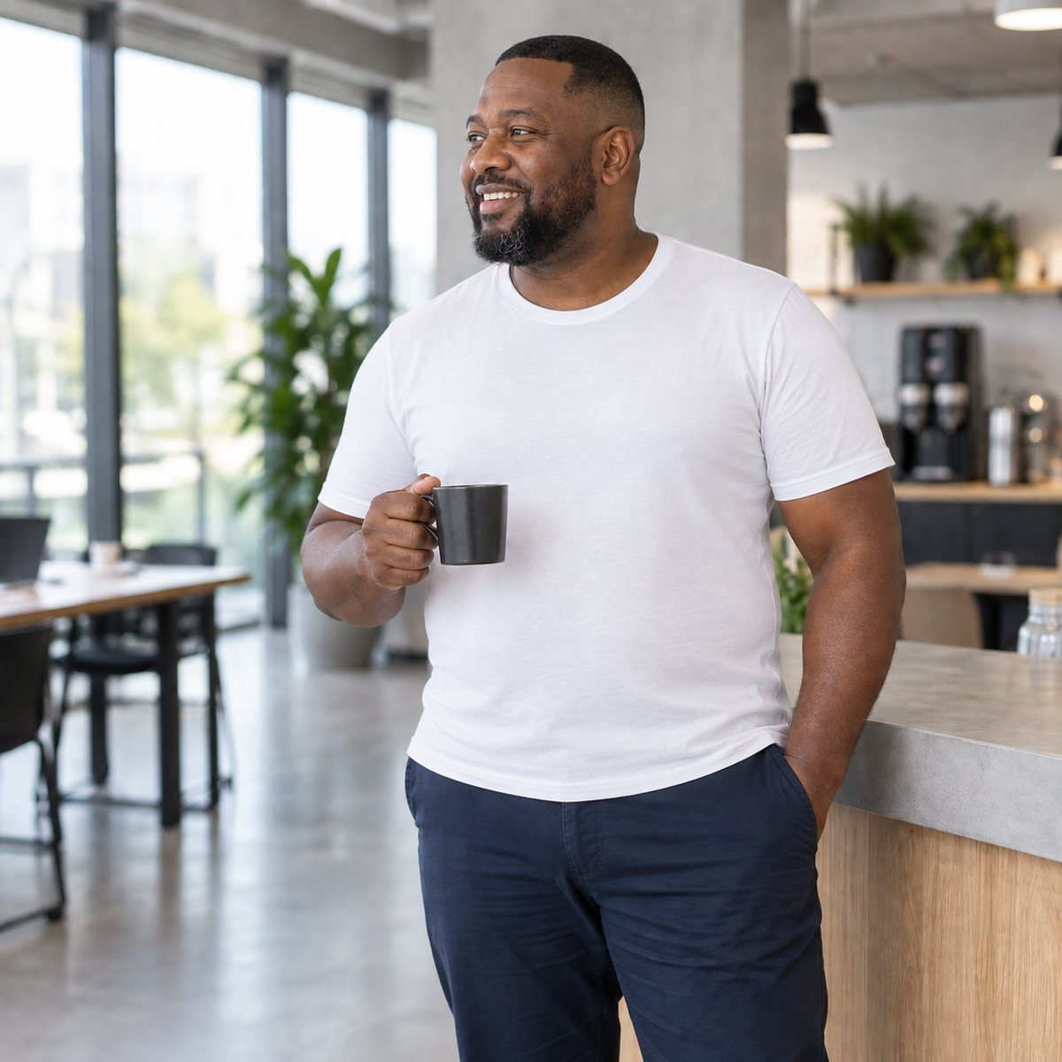 Homme riant en t-shirt blanc, un café à la main dans un bureau lumineux, buste lisse sous le coton